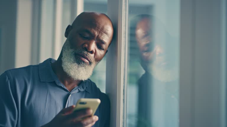 Sad, depression and senior man with smartphone by window looking outside and lonely or alone in his retirement home. Depressed, grief elderly black man waiting for text message and checking phone