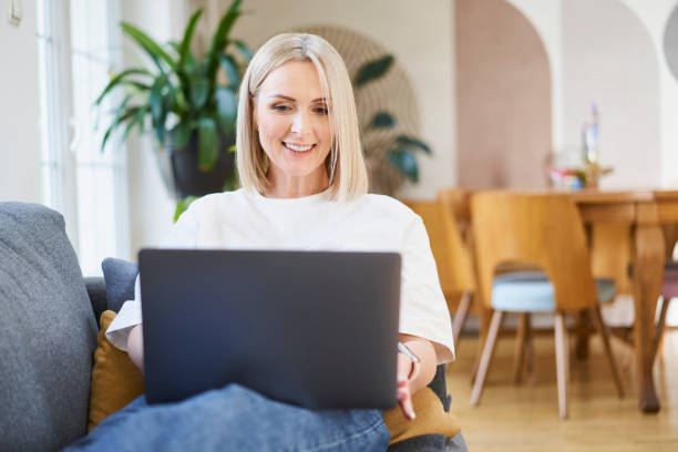 Mid adult woman at home sitting on sofa using laptop working remotely stock photo