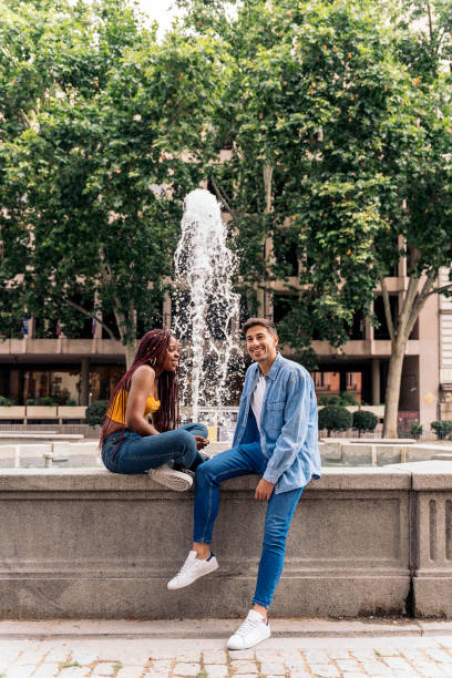 Multicultural young friends sitting next to a fountain smiling and looking at camera.