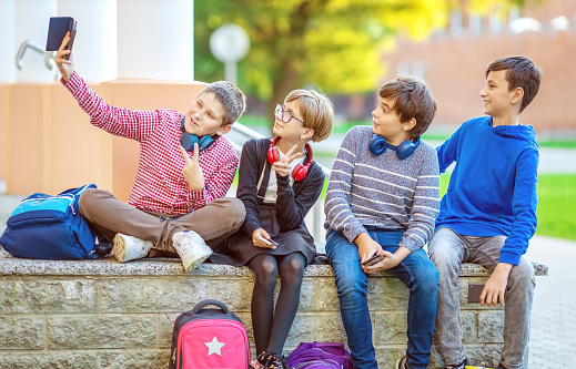 Children with rucksacks and headphones sitting on the bench in the park near the school. Kids making selfie with a phone