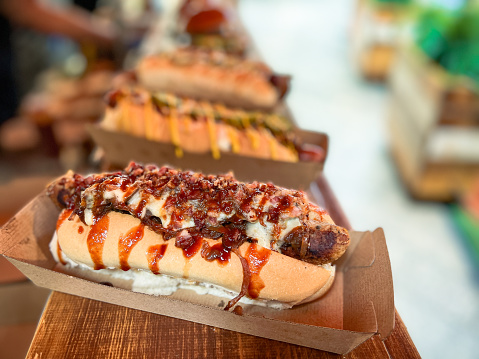 Close up image of a selection of freshly cooked hotdogs in bread rolls loaded with sauce and cheese on display at a food market.