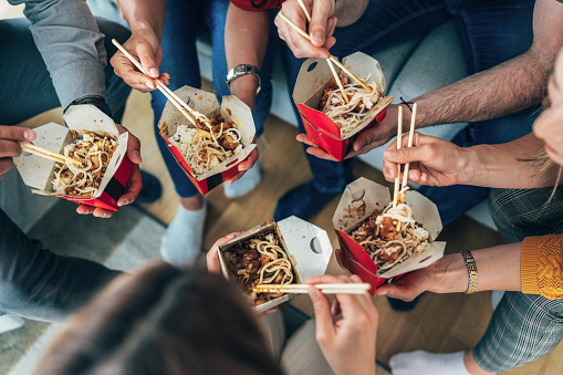 High angle view of Group of friends at home share a chinese take away food, they use chopsticks to eat. Fast food. Unrecognizable people, hands with boxes with chinese food and chopsticks.