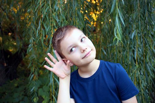 Portrait of a 9 year old boy on a background of leaves. A handsome boy in a blue t-shirt holds his hand near his ear.