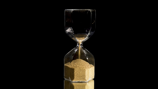 Elegant and stylish bronze transparent sandglass with trickling golden round particles. Sandglass isolated on black background, time, hurry up, last minute, no time concepts.