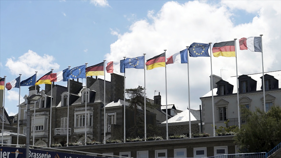 European flags swaying in the wind with beautiful cottages and blue, cloudy sky on the background. Flags of Germany, France, and European Union in front of houses.
