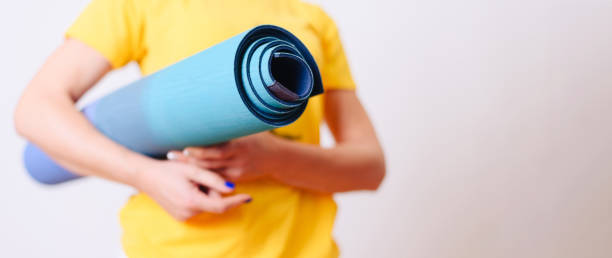 Woman dressed in a yellow T-shirt is holding a rolled up yoga mat in her hands. stock photo