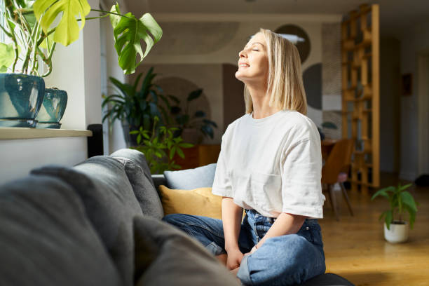 glückliche erwachsene frau, die mit geschlossenen augen auf dem sofa sitzt und helles tageslicht genießt - relaxation eyes closed women photography stock-fotos und bilder