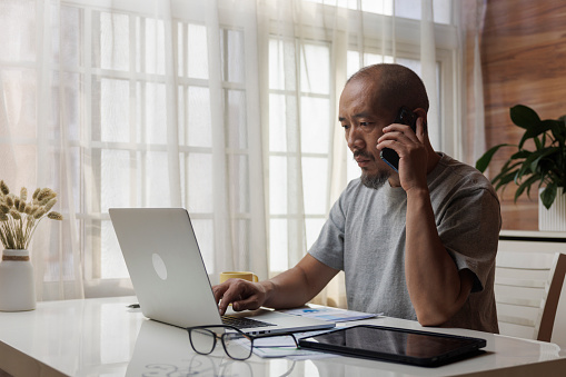 Asian middle-aged man, wearing blue shirt, sitting at the table on the balcony and working with laptop. Call the business.During the pandemic, people are working from home.