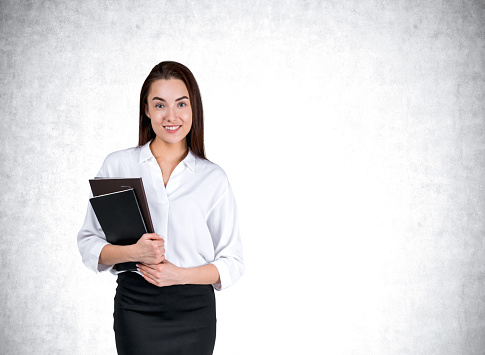 Smiling businesswoman holding journal, looking at camera with a happy smile, grey concrete wall background. Concept of learning and career. Copy space
