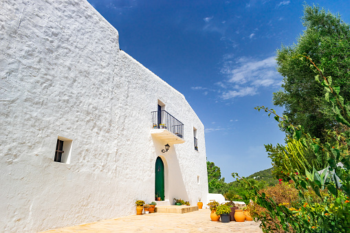 Wide-angle view of the courtyard of a typical country house located in the charming village of Sant Agustí des Vedrà, in the south-western rural outback of Ibiza, showing the essential white lime architecture of the island. A blue, bright sky, the warm light of a summer afternoon, a lovely wrought iron balcony and a picturesque lamp, ceramics and pottery overflowing with lush plants and colourful flowers. Developed from RAW.