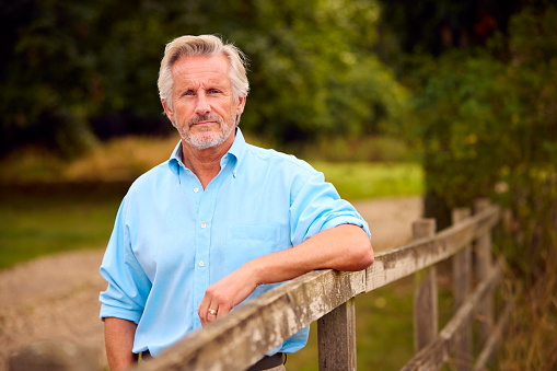 Portrait Of Casually Dressed Mature Or Senior Man Leaning On Fence On Walk In Countryside