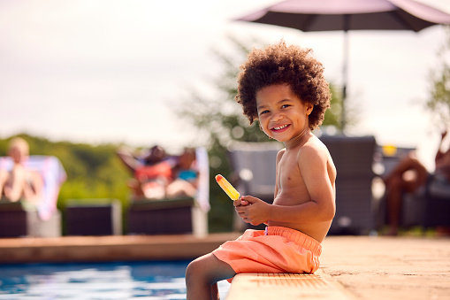 Family On Summer Holiday With Boy Eating Ice Lolly Splashing At Edge Of Swimming Pool