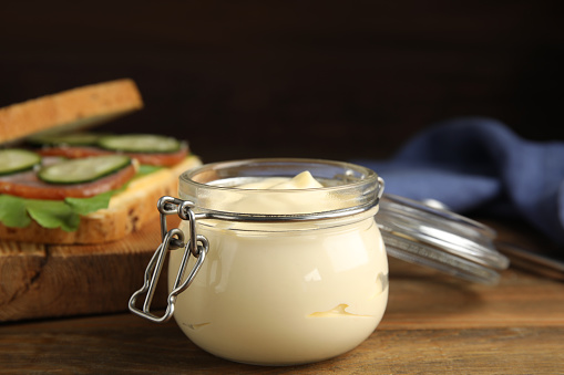 Jar of delicious mayonnaise near fresh sandwich on wooden table