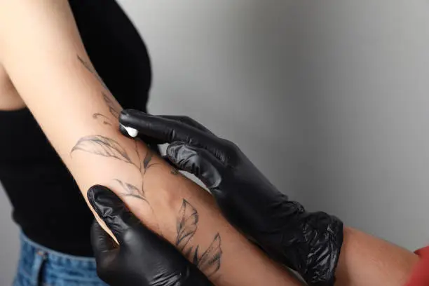 Worker in gloves applying cream on woman's arm with tattoo against light background, closeup