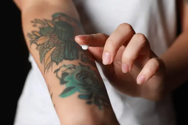 Photo of Woman applying cream on her arm with tattoos against black background, closeup