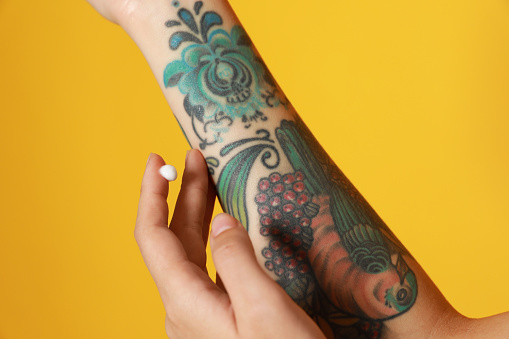 Woman applying cream on her arm with tattoos against yellow background, closeup