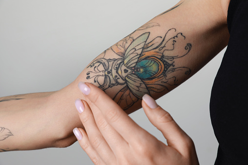 Woman applying cream on her arm with tattoos against light background, closeup