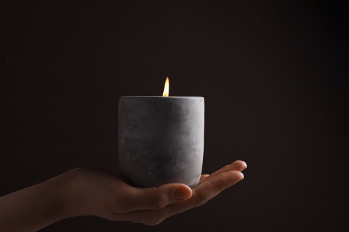 Woman with lit candle in concrete holder against dark brown background, closeup
