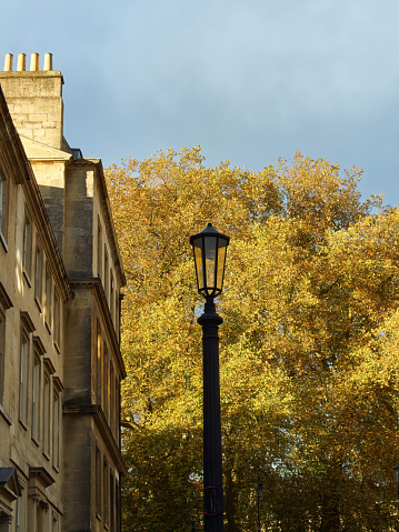 Bath in autumn - the emblematic Georgian bath stone buildings and an old ironwork lamppost in shade ahead of a tree in blazing autumn colour in bright sunlight.