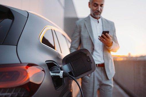 A businessman holding smartphone while charging car at electric vehicle charging station, closeup.