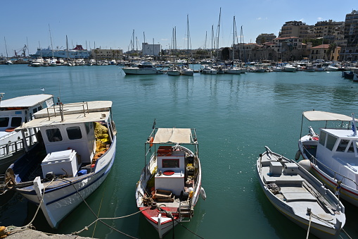 Heraklion, Greece - 22 09 2022: Old wooden fishing boats moored in port in  Heraklion near the city center.  In horizon are buildings of the town and and white private luxury yachts. und blue sky.