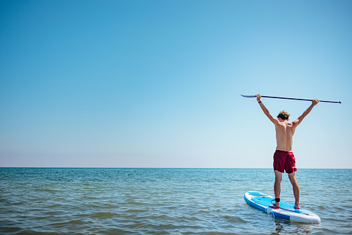 Man stands on a blue supboard and paddles. Active sport and recreation on a sunny day on the beach in the sea water.