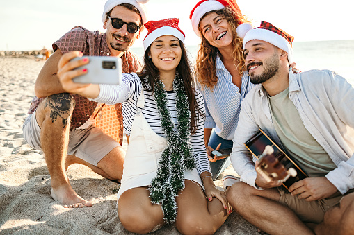 Group of people are sitting on the beach taking a selfie while wearing Santa hats