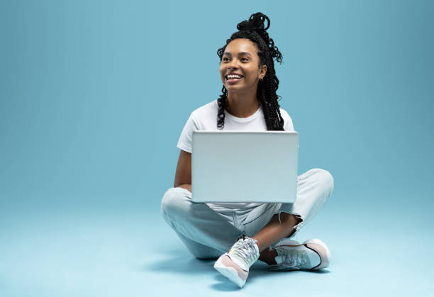 happy young woman sitting on the floor with crossed legs and using laptop on blue background. - women young adult isolated length imagens e fotografias de stock