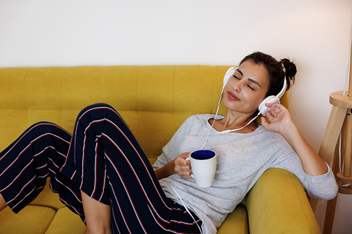 Beautiful young woman listening to music on headphones and drinking coffee while relaxing on a sofa at home