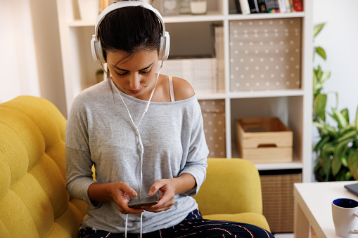 Beautiful young woman searching playlist and listening to music on headphones at home