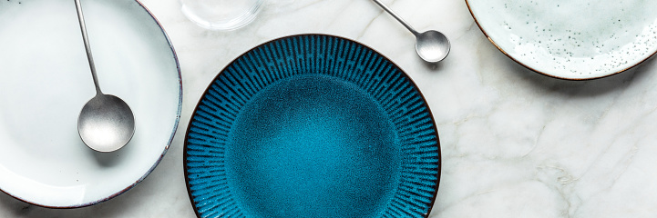 Modern tableware panorama with a vibrant blue plate and cutlery, overhead flat lay shot. Trendy dinnerware on a white marble background