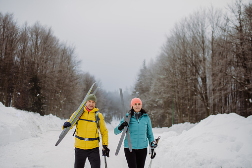 Senior couple crossing winter forest with skis on shoulders.