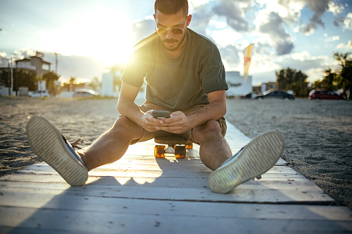 Man sits on a skateboard on the beach and uses a mobile phone