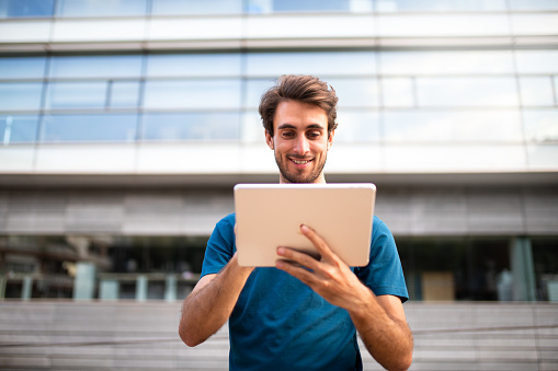 Front view of young man using digital tablet outside office buildings. Copy space. Business and technology concept.