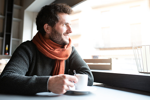 Pensive, happy young man drinking coffee in cafeteria looking out the window, enjoying early morning. Copy space. Lifestyle.