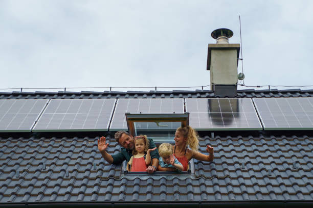 Happy family waving from skylight window in their new house with solar panels on the roof. Alternative energy, saving resources and sustainable lifestyle concept. stock photo