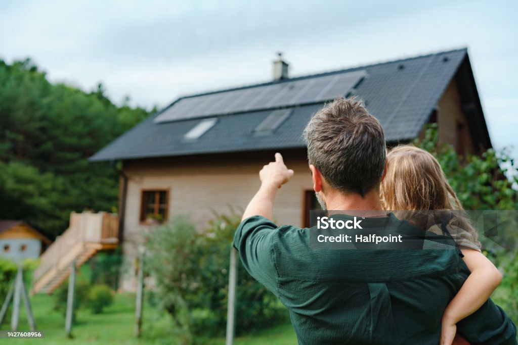 Rear view of dad holding her little girl in arms and showing at their house with installed solar panels. Alternative energy, saving resources and sustainable lifestyle concept. Rear view of dad holding her little girl in arms and showing at their house with solar panels.Alternative energy, saving resources and sustainable lifestyle concept. Solar Panel Stock Photo