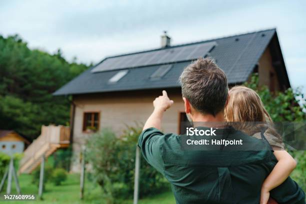 Rear View Of Dad Holding Her Little Girl In Arms And Showing At Their House With Installed Solar Panels Alternative Energy Saving Resources And Sustainable Lifestyle Concept Stock Photo - Download Image Now