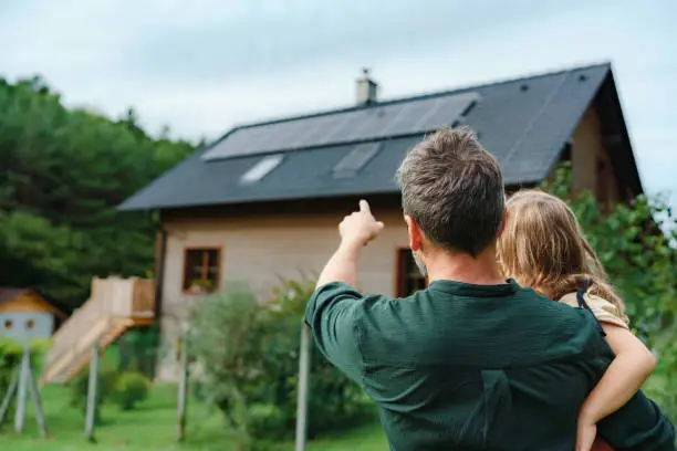 Photo of Rear view of dad holding her little girl in arms and showing at their house with installed solar panels. Alternative energy, saving resources and sustainable lifestyle concept.