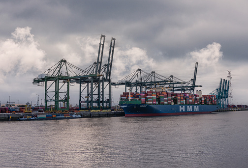 Antwerpen, Flanders, Belgium - July 10, 2022: Industry along Scheldt River. HMM loaded container vessel at Europa Terminal of port under heavy cloudscape with cranes and boxes