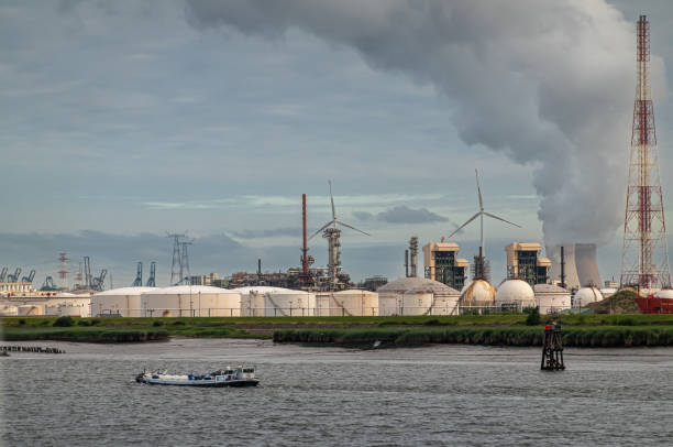 Antwerpen, Flanders, Belgium - July 10, 2022: Industry along Scheldt River. Vapor clouds out of Nuclear Reactor cooling towers behind group of white tanks at Total Olefins plant. Barge up front