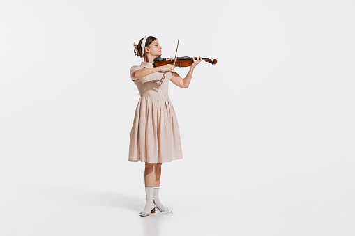 Charming beautiful young girl playing violin isolated over white studio background. Tender, lovely sound. Concept of live music, performance, retro style, creativity, artistic lifestyle