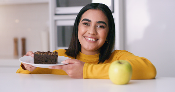 Cake, food and choice with a happy woman and dessert on a plate and an apple on the kitchen counter. Happy, snack and chocolate with a young female having a cheat day from her diet for health at home
