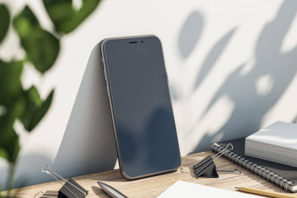 istock Perspective view on modern smartphone blank black screen with copyspace for your text or logo on wooden table with office tools and green plant on sunlit white wall background. 3D rendering, mockup 1427631627