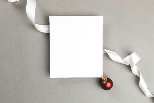 White blank card on gray background wrapped with a white ribbon. At the right bottom corner of the blank card is one red Christmas ball.