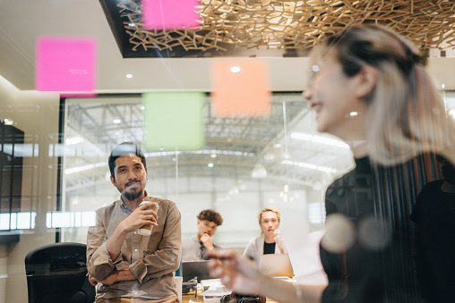 Asian diverse team of skilled young people writing their ideas for a new project on a colorful stickers glued on glass wall during a meeting in the office.