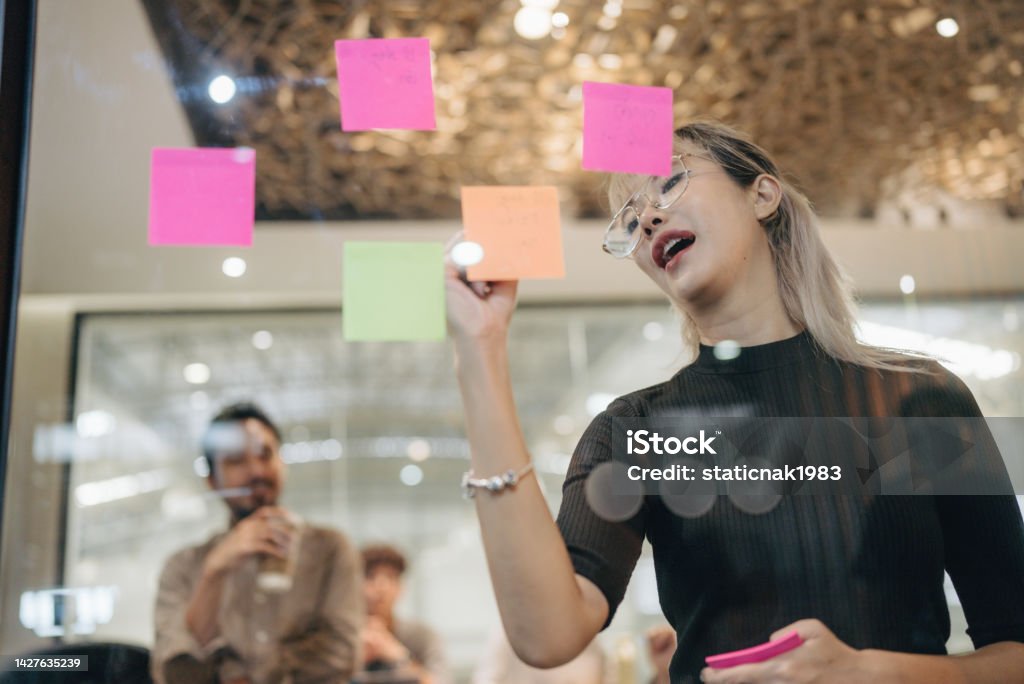 Young asian businesswomen work attack sticky notes on glass  wall in boardroom. Young asian female work on project planning board in office and having conversation with coworker friend to analyze project development . They use sticky notes posted on glass wall to make it organized . Non-Binary Gender Stock Photo