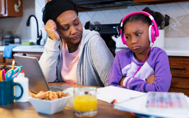 Girl and grandmother tired of doing homework An African-American girl studying at home, doing her homework on a laptop on a table in the kitchen, wearing headphones. She is pouting, arms crossed, tired of doing homework. She is almost 10 years old, an elementary student. Her grandmother is sitting next to her looking tired. grandmother real people front view head and shoulders stock pictures, royalty-free photos & images