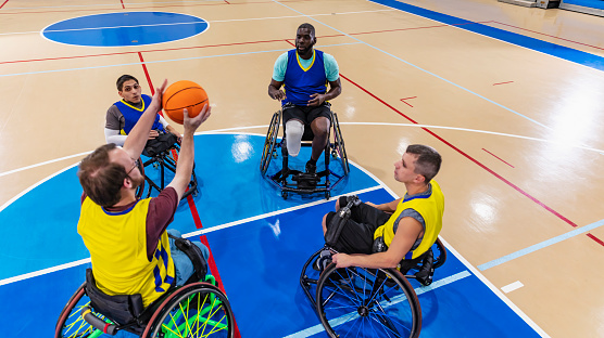 A multiracial group of four men, teammates on a wheelchair basketball team, practicing in a gym. They are sitting in a circle, passing a ball. The main focus is on the African-American man, in his 50s, who has just thrown the ball to the young man with cerebral palsy, who is reaching up to catch it.