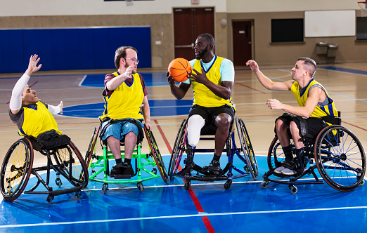 A multiracial group of four men playing on a wheelchair basketball team, practicing in a gym. The African-American man, an amputee, has the ball and all of his teammates are reaching out, trying to get him to pass it to them.